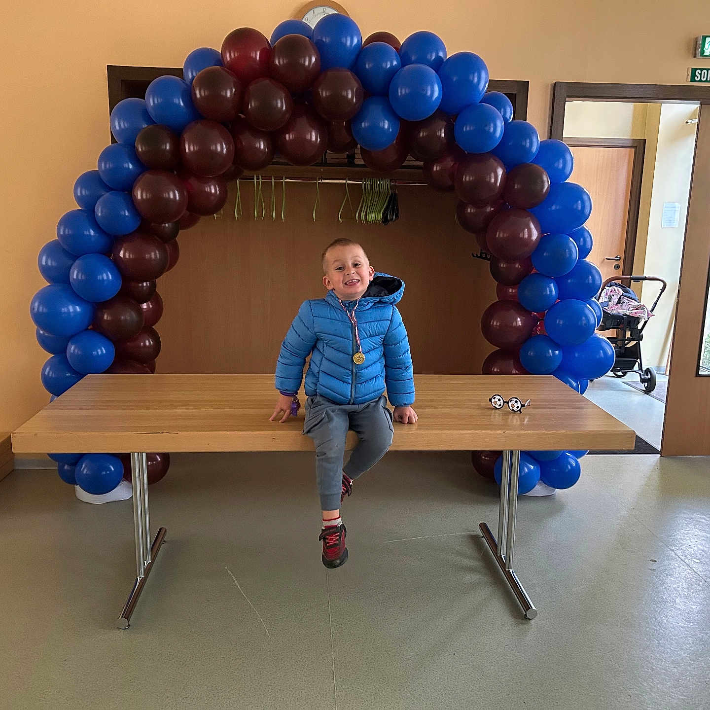 Nolann participe au concours pour gagner de l'argent avec cette photo : balloon_arch, blue_balloons, blue_jacket, boy, casual_clothing, celebration, child, door, event, exit_sign, floor, happy, indoor, maroon_balloons, medal, person, smile, stroller, table, wooden_table