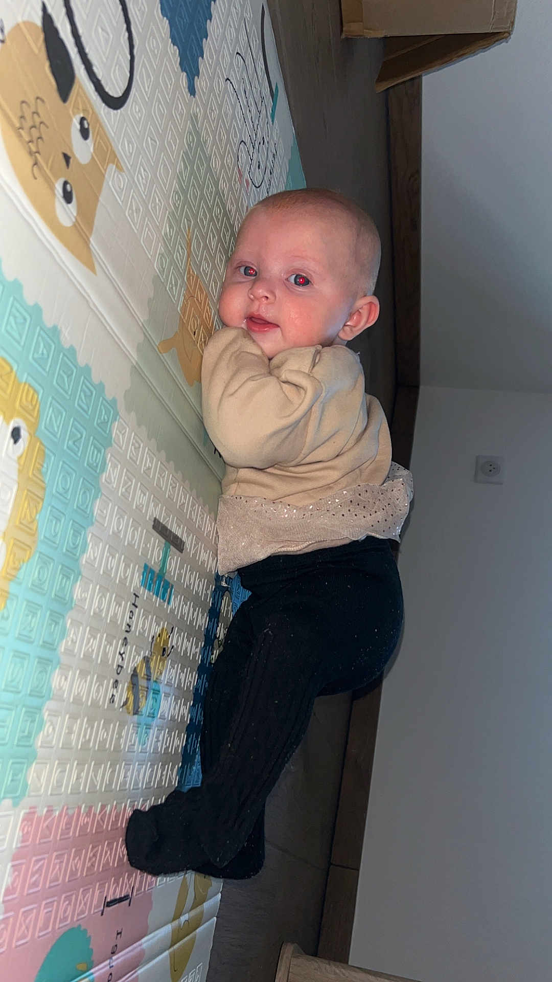 Mia participe au concours pour gagner de l'argent avec cette photo : baby, infant, play_mat, floor, indoor, child, cute, clothing, face, head, person, smile, wall, wooden_floor, furniture, socket, pants, sweater, curious, lying_down