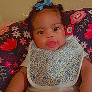 Zoe is registered to the contest to win money with this photo: baby, child, blue_bow, bib, floral_pattern, cushion, colorful, sitting, indoors, portrait, cute, infant, white_clothing, looking_at_camera, hands, legs, face, young_child, comfortable, expression