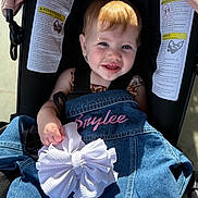 Brylee is registered to the contest to win money with this photo: toddler, child, stroller, denim_jacket, bow, smiling, blue_eyes, sunlight, earring, clothing, baby, seatbelt, outdoor, portrait, cute, happy, face, person, holding, fabric