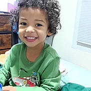 Jr is registered to the contest to win money with this photo: child, curly_hair, smiling, green_clothing, bed, blanket, indoor, window, dresser, television, cross-legged, young_child, happy, person, furniture, casual, cozy, holding_object, playful, bright