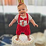 baby, birthday, cake, first_birthday, crown, red_clothing, smiling, happy, celebration, party, child, indoors, one_year_old, festive, cute, person, face, feet, white_cake, decorations
