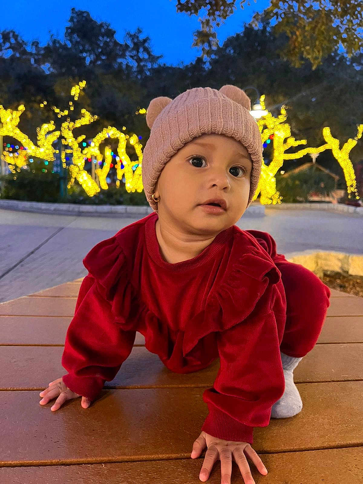 Naomi is registered to the contest to win money with this photo: toddler, child, red_clothing, knit_hat, bear_ears, crawling, wooden_surface, outdoor, night, holiday_lights, trees, face, cute, winter_wear, ears, small_hands, expression, young_child, park, evening