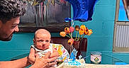 Mario is registered to the contest to win money with this photo: baby, adult, birthday_cake, balloon, number_4, table, glass_table, flower_vase, tulips, painting, indoor, celebration, person, holding, smiling, furniture, hands, wall, window, reflection