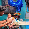baby, adult, birthday_cake, balloon, number_4, table, glass_table, flower_vase, tulips, painting, indoor, celebration, person, holding, smiling, furniture, hands, wall, window, reflection