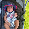 baby, child, stroller, hat, american_flag, striped_clothing, footwear, grass, outdoor, summer, infant, cute, portrait, sitting, relaxed, hand, face, expression, blue, red