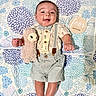 Mario is registered to the contest to win money with this photo: baby, infant, child, stuffed_animal, llama, quilt, floral_pattern, clothing, suspenders, bowtie, smiling, lying_down, wooden_sign, months_marker, cute, soft_toy, portrait, happy, indoors, person
