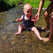 Kensley is registered to the contest to win money with this photo: baby, child, water, creek, nature, sunlight, swimsuit, adult, hand, rocks, outdoor, grass, stream, sitting, barefoot, summer, greenery, shallow_water, person, playing