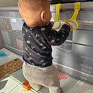 Lou a rejoint le concours — aidez-le/la à gagner de superbes lots ! baby, baby_clothes, back_view, child, childhood, floor_mat, home, indoor, infant, padded_floor, plastic_rings, play_area, playpen, puzzle, standing, sunlight, toddler, toys, yellow_rings, young_child