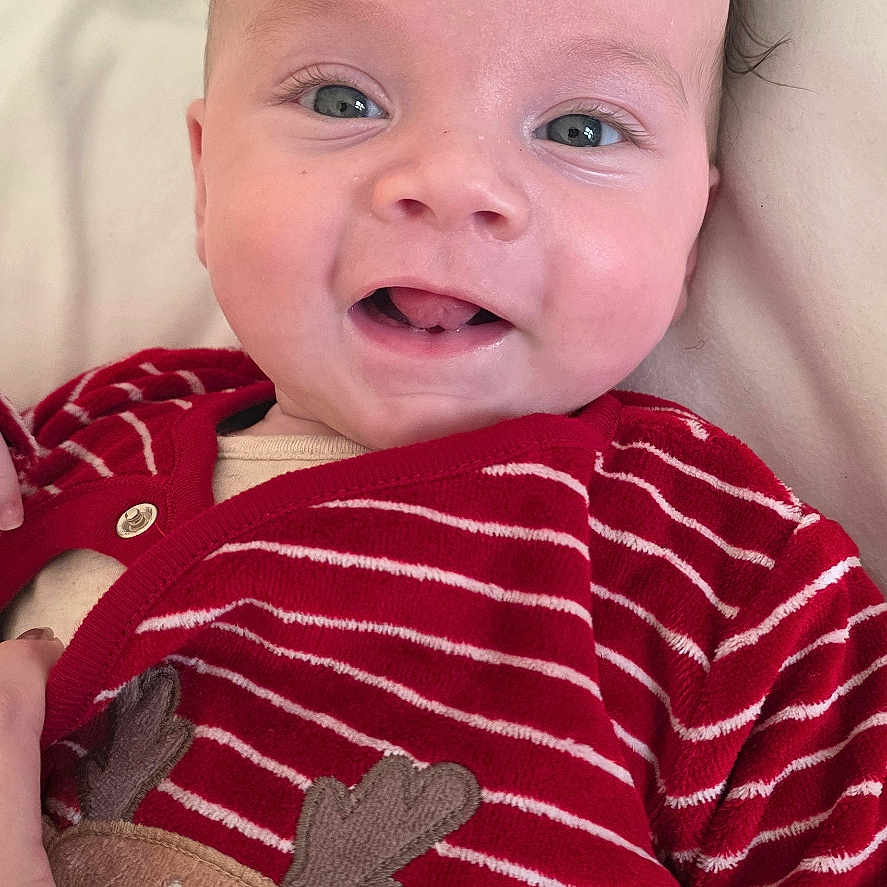 Jake is registered to the contest to win money with this photo: baby, child, closeup, cozy, cute, expression, eyes, face, hand, happy, infant, lying_down, mouth, newborn, red_clothing, reindeer_applique, skin, smile, soft_surface, striped
