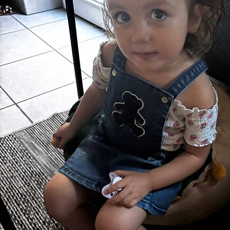 Sophie participe au concours pour gagner de l'argent avec cette photo : carpet, child, curious, cushion, denim_dress, floor_tiles, footwear, girl, hair_clip, hand_holding_object, indoor, looking_up, pacifier, patterned_clothing, ponytail, sandals, seated, short_sleeves, toddler, young_child