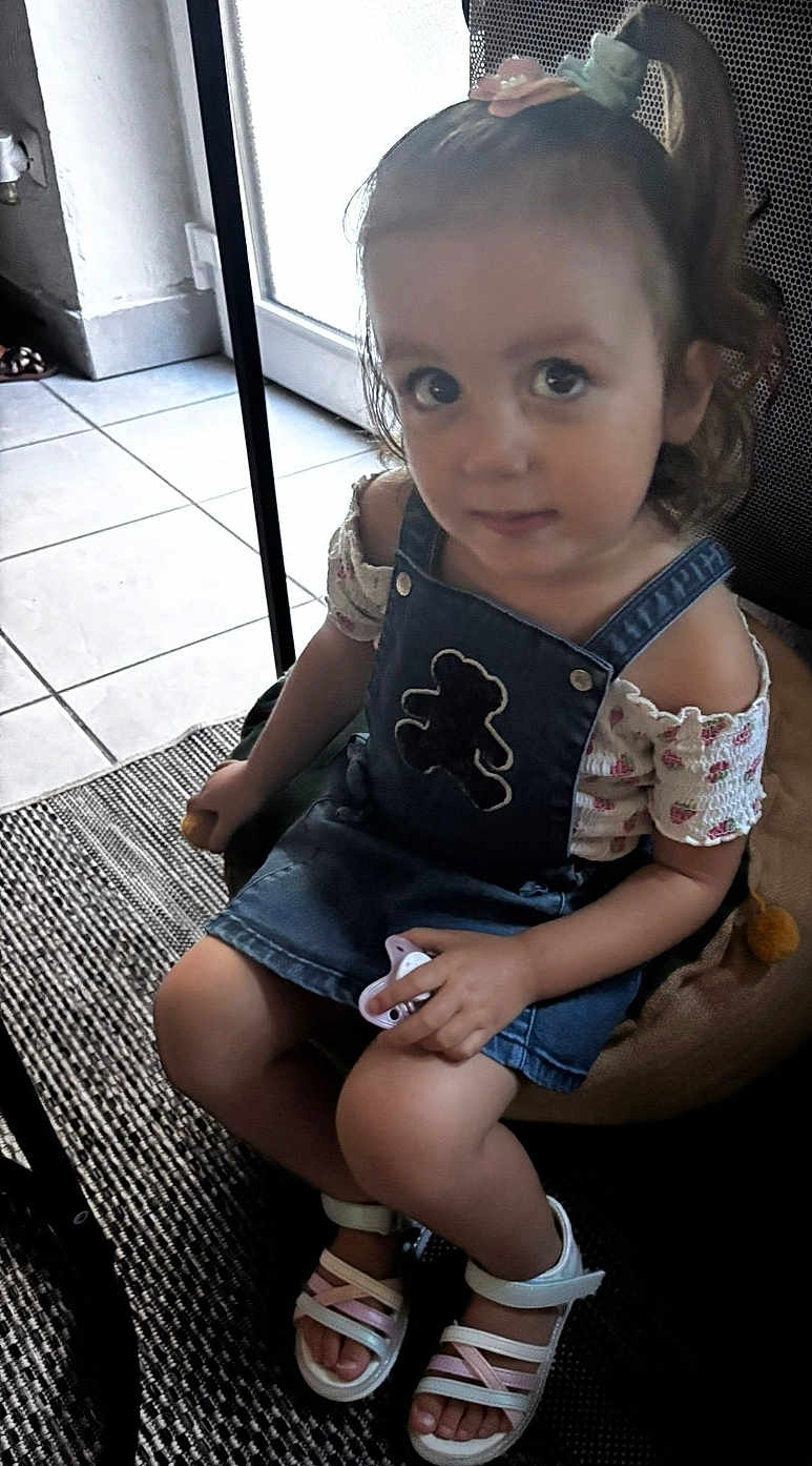 Sophie participe au concours pour gagner de l'argent avec cette photo : toddler, child, girl, indoor, denim_dress, pacifier, hair_clip, ponytail, footwear, sandals, floor_tiles, cushion, carpet, seated, curious, looking_up, short_sleeves, patterned_clothing, hand_holding_object, young_child