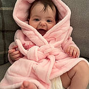 Isabella is registered to the contest to win money with this photo: baby, infant, pink_robe, couch, smiling, happy, child, diaper, furniture, cute, hood, indoor, soft_texture, sitting, foot, hand, cozy, plush, face, portrait