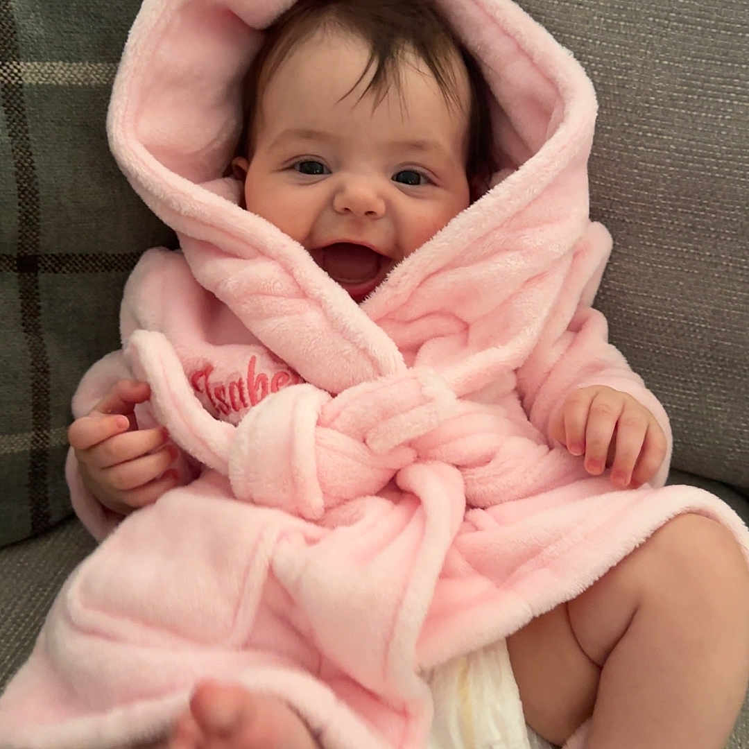 Isabella is registered to the contest to win money with this photo: baby, child, couch, cozy, cute, diaper, face, foot, furniture, hand, happy, hood, indoor, infant, pink_robe, plush, portrait, sitting, smiling, soft_texture
