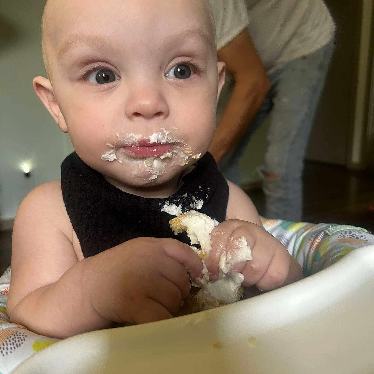 Miles joined the competition — help win amazing prizes! adult, baby, black_bib, cake, child, closeup, curious, eating, expression, face, fingers, food, frosting, hands, high_chair, indoor, messy, mouth, person, young_child