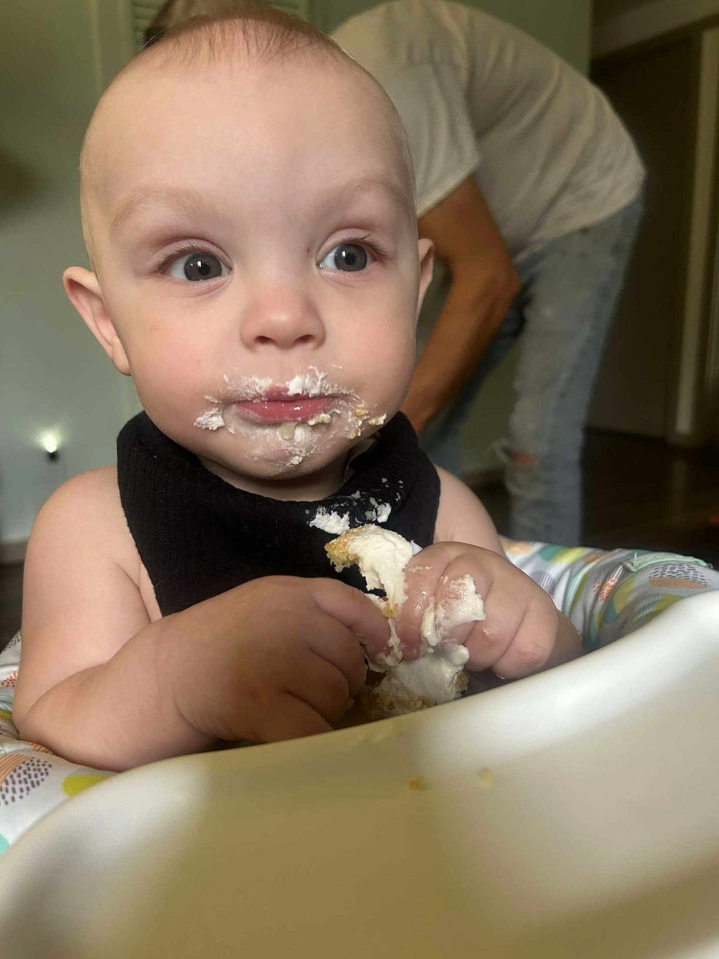 Miles joined the competition — help win amazing prizes! baby, child, face, frosting, food, high_chair, messy, hands, indoor, person, adult, curious, cake, eating, closeup, young_child, black_bib, fingers, mouth, expression
