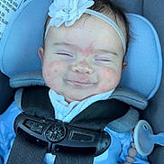 Addison is registered to the contest to win money with this photo: baby, child, car_seat, maxi_cosi, headband, flower, pacifier, smile, closed_eyes, blue_clothing, straps, safety_harness, infant, cute, indoors, person, seat, comfort, resting, happy