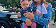 Addison is registered to the contest to win money with this photo: baby, black_vehicle, car, casual, child, crowd, daylight, denim_jacket, headband, holding, nature, outdoor, park, person, pink_clothing, portrait, sidewalk, smile, trees, woman