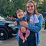 woman, baby, child, denim_jacket, pink_clothing, headband, outdoor, car, black_vehicle, trees, crowd, sidewalk, person, holding, smile, nature, daylight, casual, portrait, park