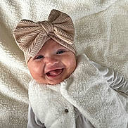 Hélia a rejoint le concours — aidez-le/la à gagner de superbes lots ! baby, smiling, headband, bow, vest, white_clothing, lying_down, blanket, soft_texture, happy, infant, child, cute, portrait, indoor, closeup, face, warm_clothing, expression, adorable