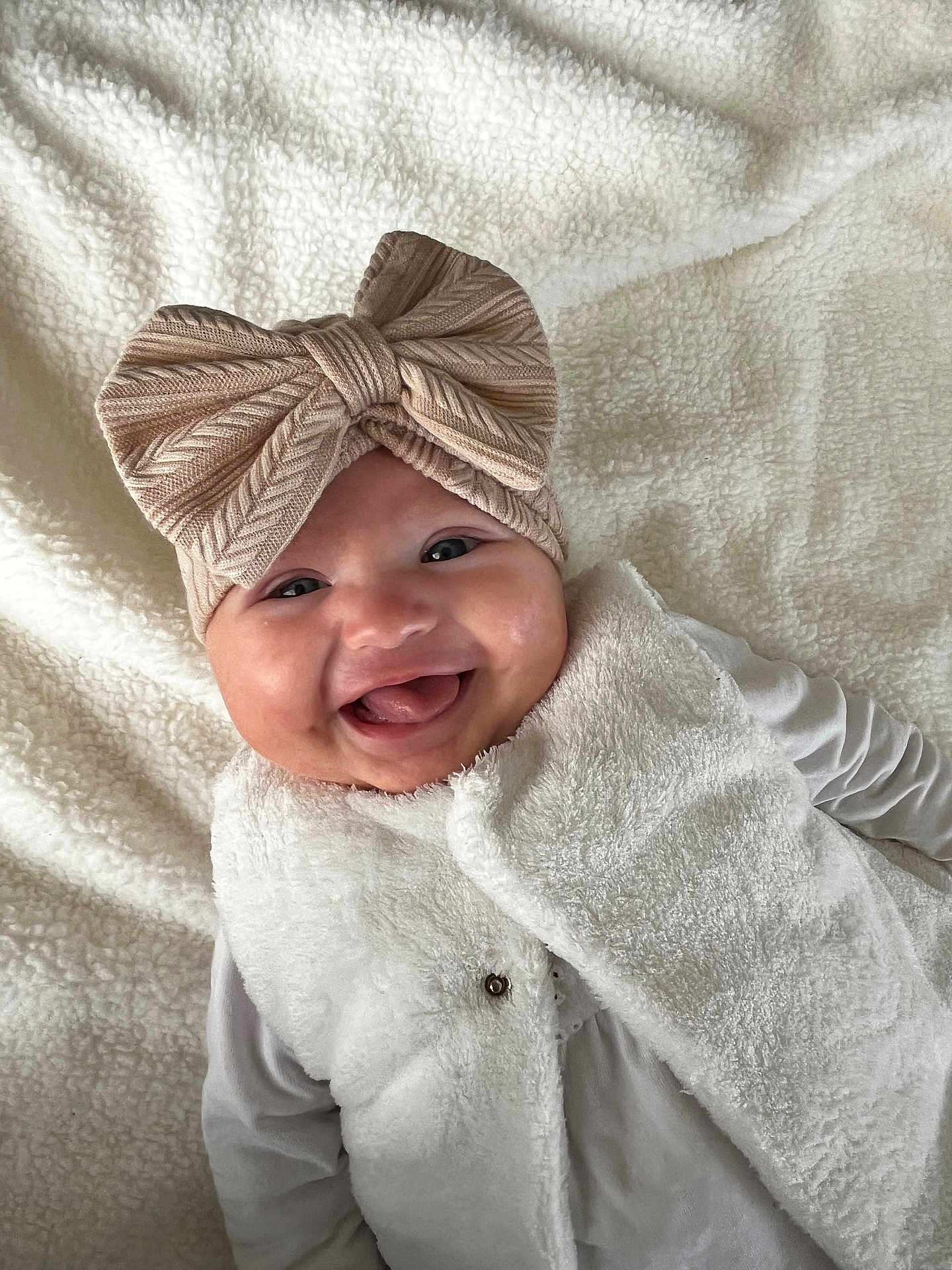 Hélia a rejoint le concours — aidez-le/la à gagner de superbes lots ! baby, smiling, headband, bow, vest, white_clothing, lying_down, blanket, soft_texture, happy, infant, child, cute, portrait, indoor, closeup, face, warm_clothing, expression, adorable