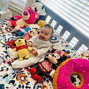 Andrea is registered to the contest to win money with this photo: baby, smiling, plush_toy, bed, colorful, window_blinds, winnie_the_pooh, mickey_mouse, minnie_mouse, stuffed_animal, pink_donut, toy_collection, happy, indoors, child, soft_lighting, floral_bedspread, cute, sitting, playful