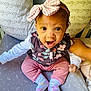 baby, child, infant, pink_bow, headband, socks, floral_vest, pink_pants, cushion, hand, indoor, surprised_expression, seated, patterned_bedding, cute, portrait, person, young_child, cozy, supporting_hand