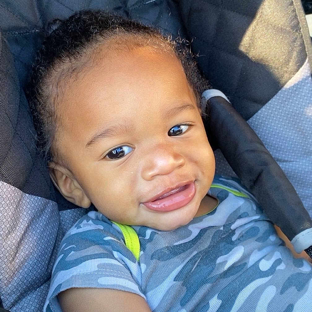 Namir joined the competition — help win amazing prizes! toddler, child, smile, curly_hair, stroller, blue_shirt, camo_pattern, seat, sunlight, happy, portrait, baby, infant, outdoor, casual_clothing, closeup, person, young_child, relaxing, daylight