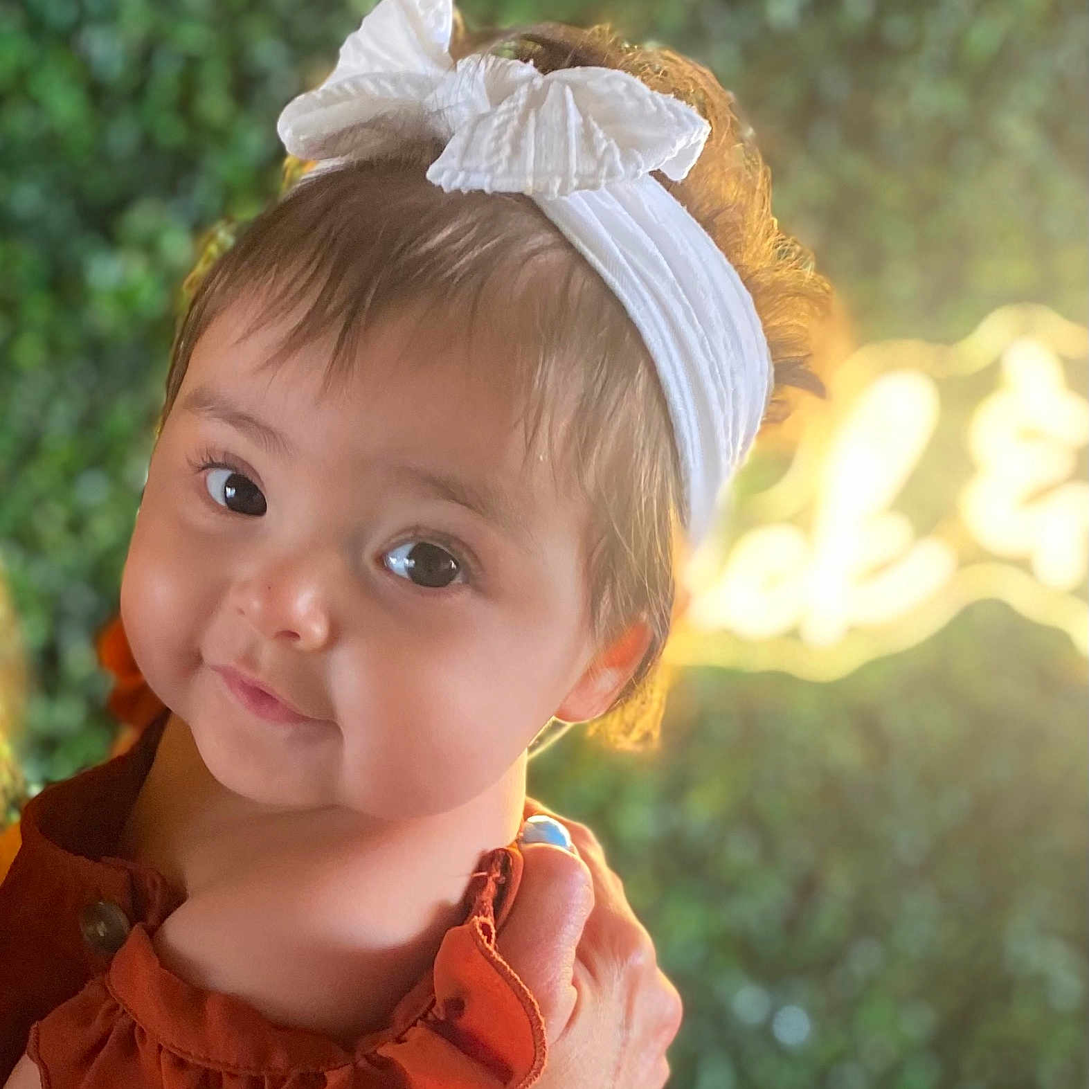Octavia is registered to the contest to win money with this photo: baby, blurred_background, bow, child, clothing, cute, expression, face, greenery, hand, headband, holding, infant, outdoor, person, portrait, skin, smile, sunlight, warm_light