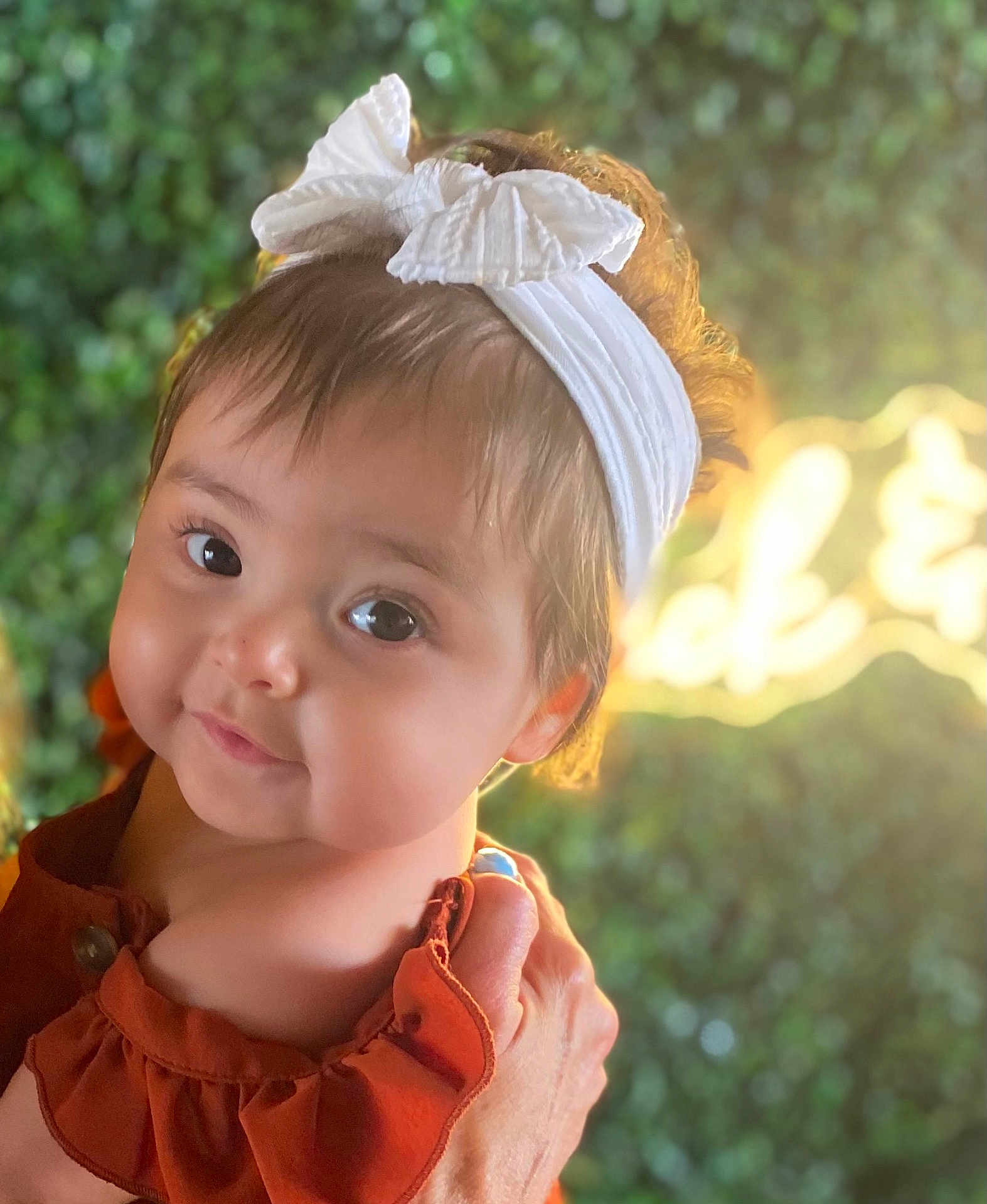 Octavia is registered to the contest to win money with this photo: baby, child, headband, bow, face, portrait, skin, hand, clothing, person, cute, smile, outdoor, greenery, blurred_background, sunlight, warm_light, infant, expression, holding