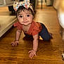 baby, child, crawling, headband, flower_pattern, orange_top, wooden_floor, indoors, furniture, curious, smiling, cute, portrait, person, flooring, headwear, young_child, home, floor, candid