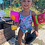 toddler, child, smiling, swimsuit, floaties, corn, shoe, barefoot, outdoor, sunny, patio, fence, plants, greenery, summer, playful, happy, food, water, daylight