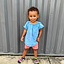 toddler, child, smiling, standing, blue_top, pink_shorts, colorful_socks, concrete, gray_wall, outdoor, curly_hair, casual_clothing, person, footwear, fun_socks, playful, young_child, portrait, daylight, urban