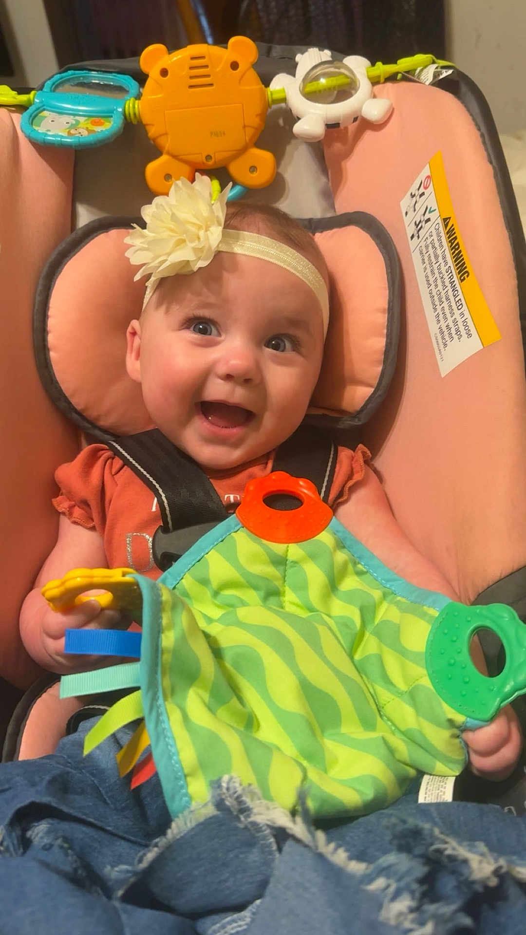 Malayha is registered to the contest to win money with this photo: baby, infant, car_seat, headband, flower, toy, colorful, green, yellow, pink, blue, clothing, happy, smiling, child, cute, face, hands, fabric, seatbelt