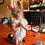 child, bunny_mask, sunglasses, kitten, floor, wood_floor, tshirt, diaper, living_room, television, furniture, clock, stockings, stacked_clothes, toy, pet, cute, playful, indoors, fun
