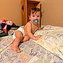 bare_chest, bed, blanket, brown_wall, child, curly_hair, cute, diaper, dinosaur_sheets, foot, furniture, hand, indoor, infant, looking_at_camera, pacifier, playroom, toddler, toy, wooden_shelf