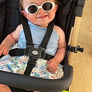 Maïa participe au concours pour gagner de l'argent avec cette photo : accessory, baby, barefoot, black_stroller, blue_bow, child, clothing, cute, fashion, floral_skirt, happy, indoor, infant, person, playful, portrait, seatbelt, smile, stroller, sunglasses