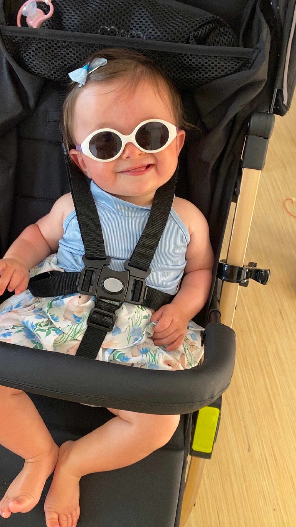 Maïa participe au concours pour gagner de l'argent avec cette photo : accessory, baby, barefoot, black_stroller, blue_bow, child, clothing, cute, fashion, floral_skirt, happy, indoor, infant, person, playful, portrait, seatbelt, smile, stroller, sunglasses