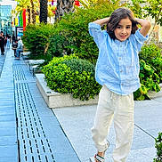 Ilian participe au concours pour gagner de l'argent avec cette photo : child, urban, sidewalk, plants, greenery, palm_trees, blue_shirt, white_pants, sandals, confident, smiling, outdoor, daylight, people, modern, casual_clothing, trees, bench, shopping_area, relaxing
