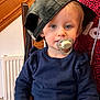 toddler, child, pacifier, hat, blue_sweater, indoor, chair, wooden_furniture, radiator, person, face, headwear, casual_clothing, cute, baby, sitting, young_child, portrait, light_skin, blonde_hair