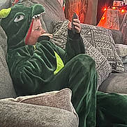 Adian is registered to the contest to win money with this photo: casual, child, costume, couch, cozy, cushion, dinosaur_onesie, face, focused, ghost_decoration, green_clothing, halloween_decorations, holiday, home, indoor, lights, person, relaxed, sitting, smartphone