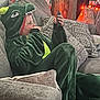 casual, child, costume, couch, cozy, cushion, dinosaur_onesie, face, focused, ghost_decoration, green_clothing, halloween_decorations, holiday, home, indoor, lights, person, relaxed, sitting, smartphone