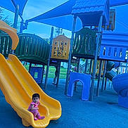 Tasneem joined the competition — help win amazing prizes! toddler, child, playground, slide, yellow_slide, blue_floor, play_equipment, tic_tac_toe, outdoor, shade_sails, climbing_structure, park, recreation, greenery, blue_sky, people_in_background, casual_clothing, sitting, curious, daytime