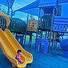 toddler, child, playground, slide, yellow_slide, blue_floor, play_equipment, tic_tac_toe, outdoor, shade_sails, climbing_structure, park, recreation, greenery, blue_sky, people_in_background, casual_clothing, sitting, curious, daytime
