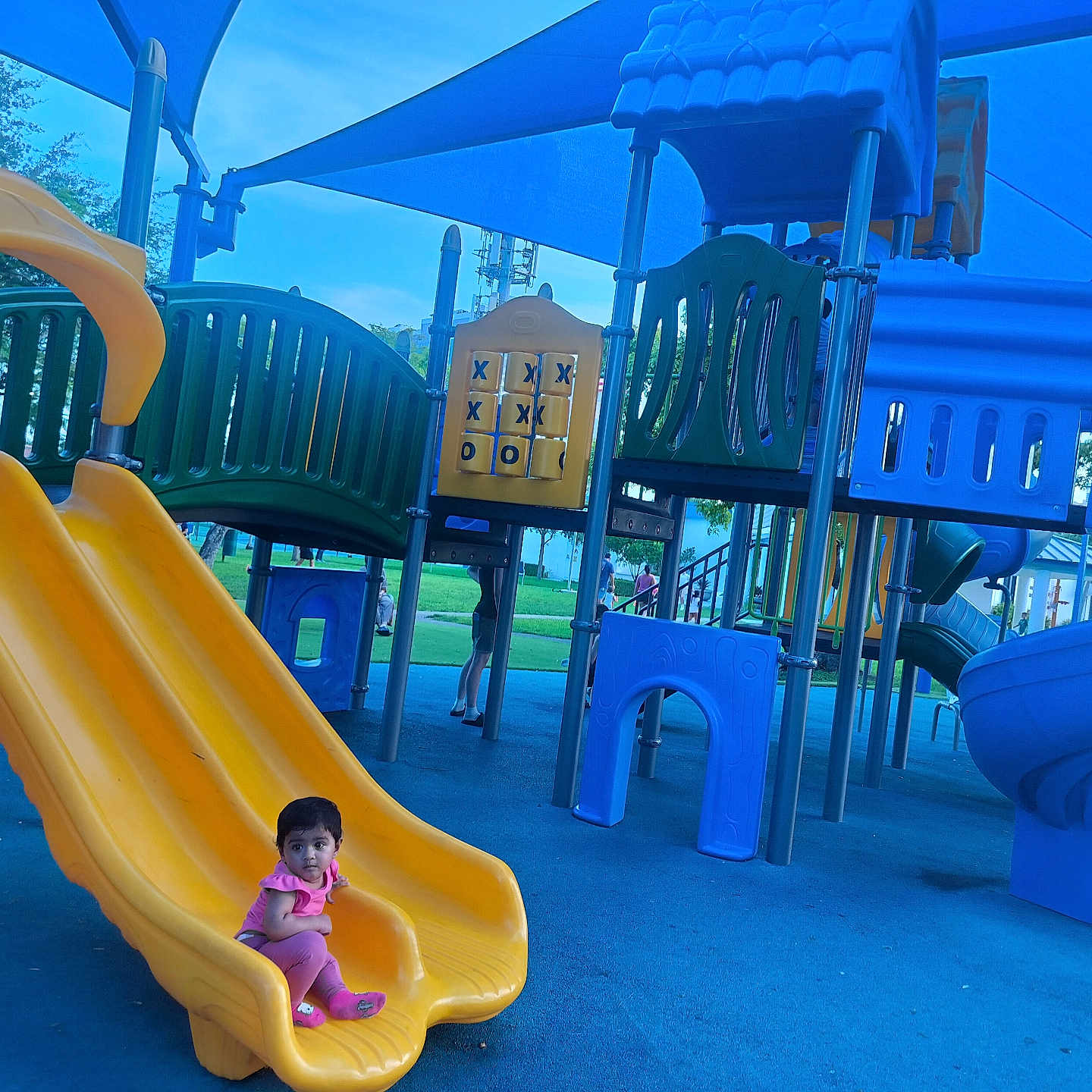 Tasneem joined the competition — help win amazing prizes! toddler, child, playground, slide, yellow_slide, blue_floor, play_equipment, tic_tac_toe, outdoor, shade_sails, climbing_structure, park, recreation, greenery, blue_sky, people_in_background, casual_clothing, sitting, curious, daytime