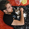 man, cat, bean_bag, blanket, glasses, indoor, floor, snack_bag, tabby_cat, relaxing, petting, casual, black_shirt, orange, home, carpet, human, comfort, resting