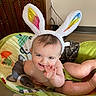 baby, bunny_ears, baby_seat, smiling, child, indoor, blanket, wooden_furniture, playful, cute, infant, face, hand, legs, colorful, comfort, home, person, cheerful, soft