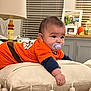 baby, pacifier, orange_shirt, cushion, tassels, living_room, lamp, framed_photos, decor, window_blinds, child, indoor, curious, toddler, person, face, hand, furniture, home, cozy