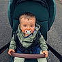 baby, pacifier, stroller, child, outdoor, pavement, green_jacket, light_pants, white_socks, curious, seated, baby_face, infant, hands, safety_straps, hooded_jacket, cute, young_child, baby_clothing, daylight