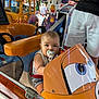 toddler, pacifier, amusement_ride, toy_vehicle, cartoon_vehicle, colorful, child, outdoor, person, cow_figure, summer, daylight, trees, shorts, seatbelt, playground, fun, family, cute, baby